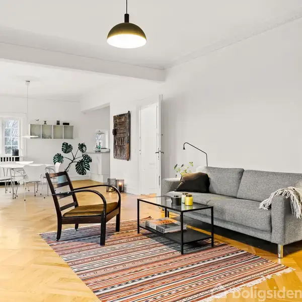 Grå sofa står ved siden af et sofabord og en stol, placeret på et stribet tæppe i en lys stue. I baggrunden er der et køkkenområde med spisebord.