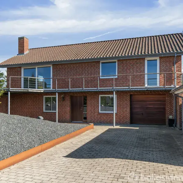 Murstenshus med garage og carport. Huset har skråtag med tegl og to vinduer. Garagen har en hvid dør og et nummer "25" ved siden af. Flisebelagt indkørsel ved huset.