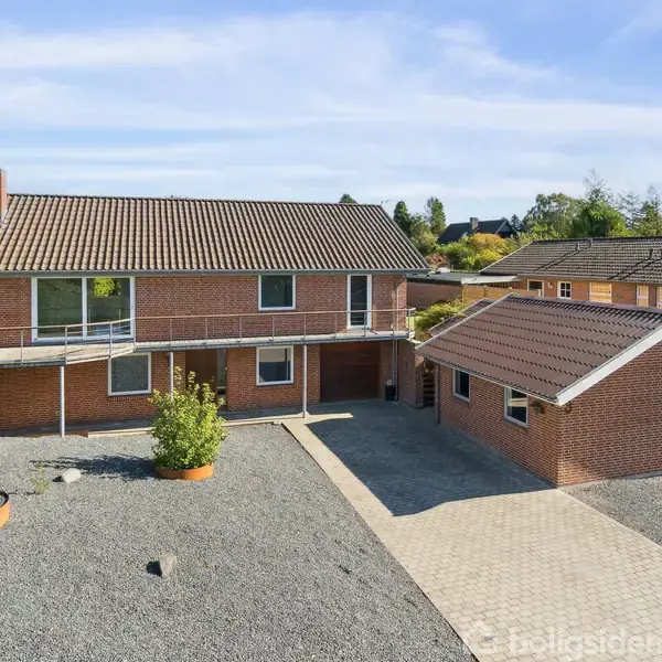 Røde murstensbygninger med flisebelagt gangsti strækker sig mellem dem, hvor en skråning af sten og en parkeringsplads med en bil ses i baggrunden under en klar blå himmel.