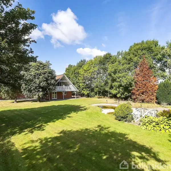 En velholdt grøn græsplæne med et lille haveanlæg inklusive en dam og bro er omgivet af træer. I baggrunden ses et rødt hus under en blå himmel med skyer.