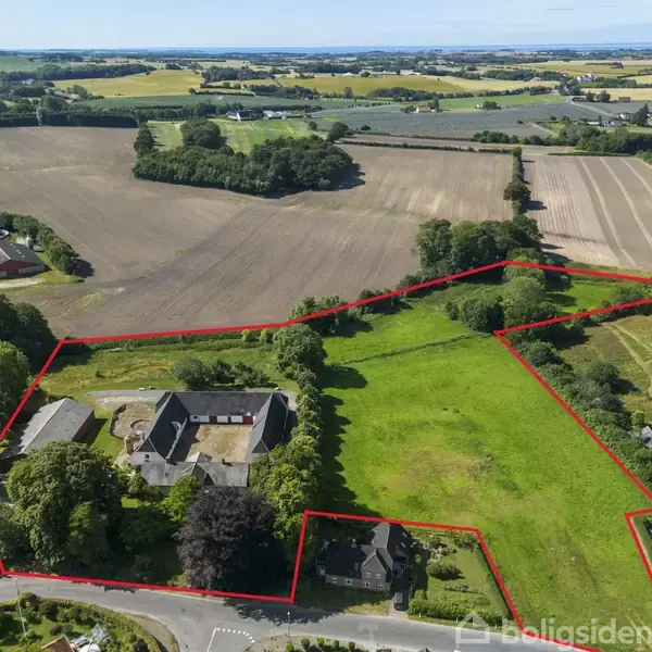 Landområde indrammet med rød linje spreder sig over marker og bygninger, omgivet af grønne marker og en landevej. Adskillige træer og åbne marker ses i baggrundens landskab.