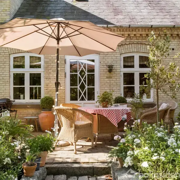 Parasol skygger udendørs spiseplads med fletstole og rødtternet dug. Omgivet af blomstrende planter foran et beige murstenshus med hvide vinduer og en terrasse.