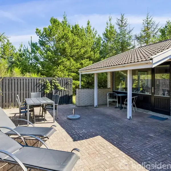 Liggestole står på en flisebelagt terrasse ved siden af et halvtag tilknyttet et hus. Omringet af høje grønne træer og sorte hegn, skaber privat og fredelig atmosfære.