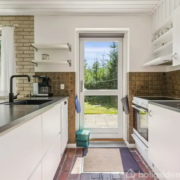 Et køkken med hvide skabe og sort bordplade åbner ud mod en terrasse gennem en glasdør. Udenfor ses en grøn have. En kaffemaskine står på bordet.