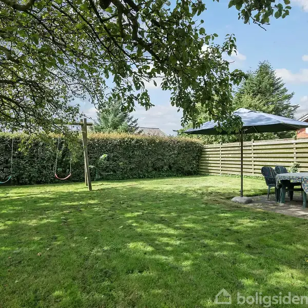 Et græsbevokset haveområde med gynger hængende fra en metalramme. Ved siden af står et bord med stole under en parasol. Hækken og et hus danner baggrunden.