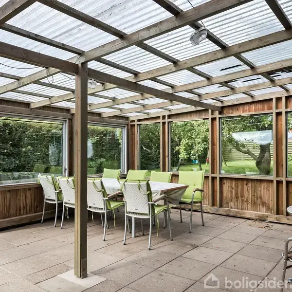 En overdækket terrasse har et langbord med grønne stole. Det hele er omgivet af store vinduer og en frodig have ses udenfor.