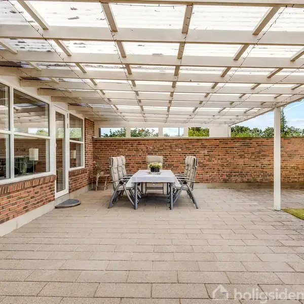 Et udendørs terrasseområde med et spisebord og seks stole, står under en glasoverdækning ved siden af en murstensvæg. Grøn græsplæne kan ses til højre.