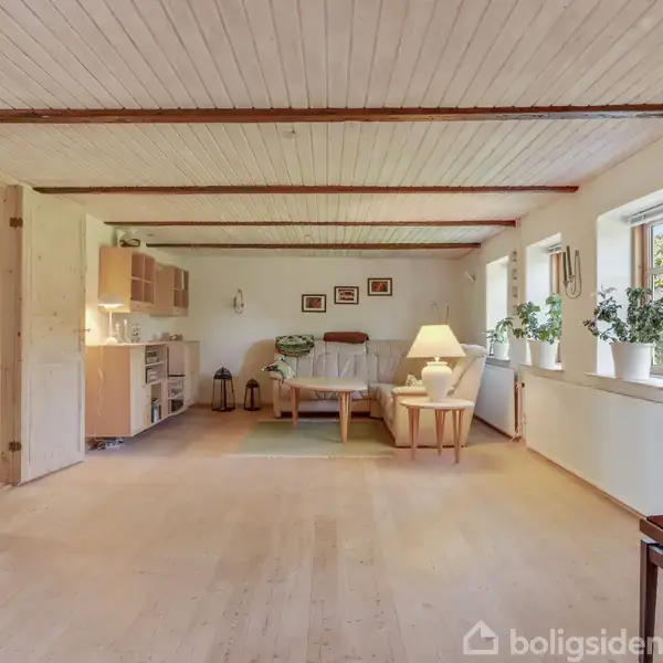 Stue med træloft, sofaområde og planter på vindueskarmen. Et elektrisk klaver står ved siden af, og en bordlampe lyser hjørnet op. Væggene har billeder og skabene er lyse.