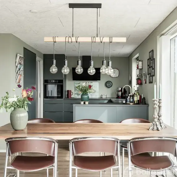 Et moderne køkken med et træbord og seks stole. Bordet har en vase med blomster og stearinlys. Bagved er et køkkenområde med skabe, et komfur og hængende lamper.