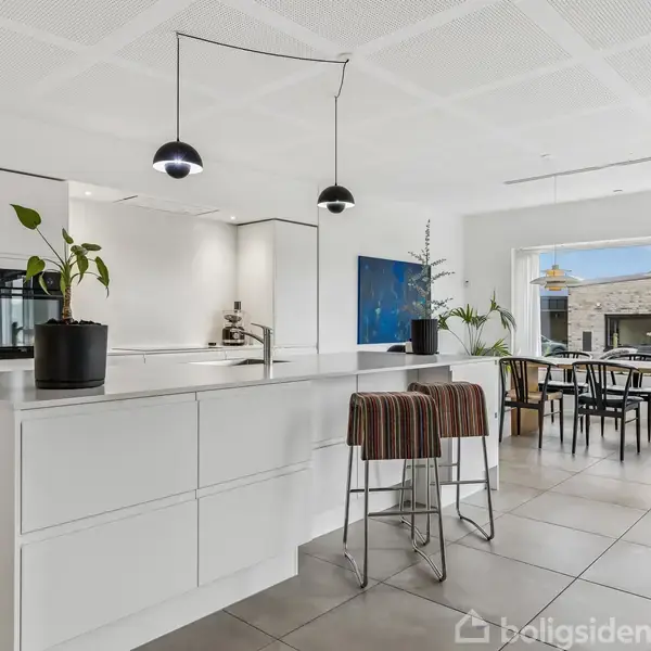 Køkkenø med potteplanter og høje stole står i et moderne, lyst køkken. I baggrunden ses et spisebord med stole og store vinduer med udsigt til en terrasse.