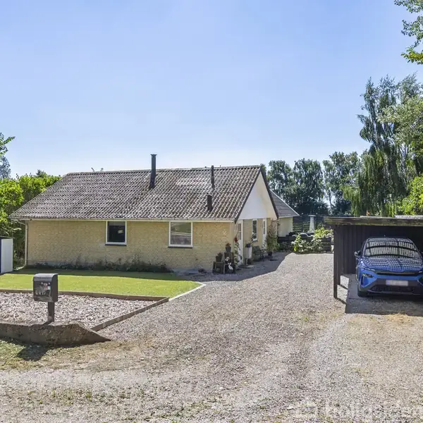 Huset står stille i et åbent område omgivet af træer, med en carport til to biler ved siden af, grusvej foran og en lille græsplæne foran.