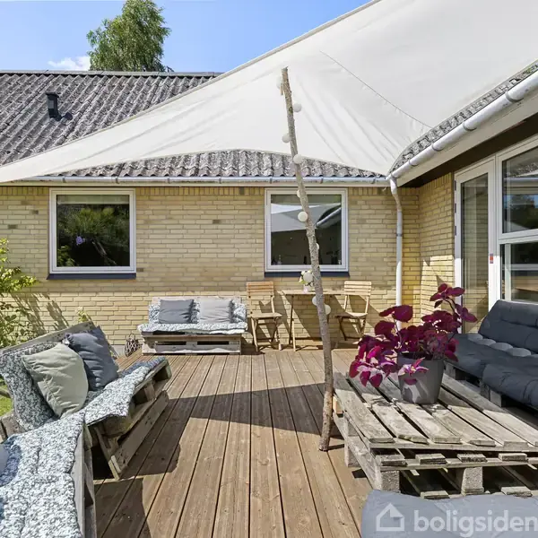 Terrasse med loungemøbler i grå nuancer lavet af paller, overdækket med et sejl. Murstenshus med vinduer i baggrunden, omgivet af planter og klart solskinsvejr.