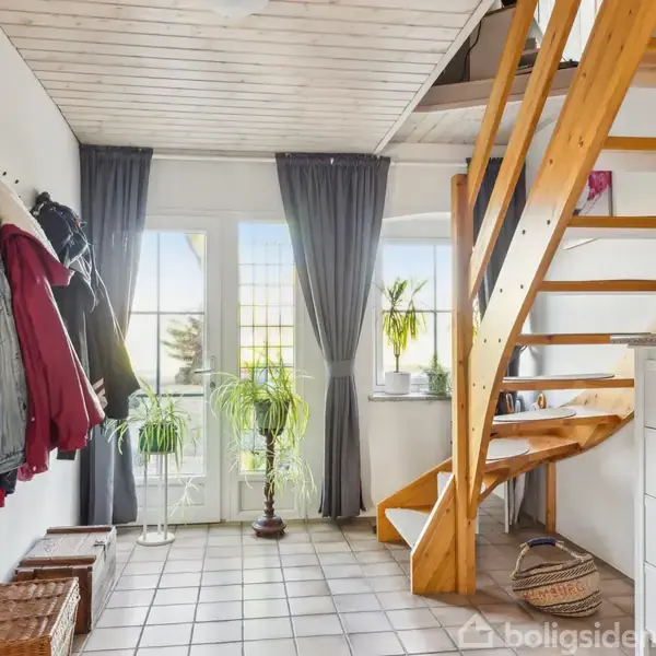 Trappe med trætrin fører opad i en lys entré med flisegulv og hvide vægge. Til venstre hænger jakker, og lyset strømmer ind igennem store vinduer med grå gardiner.
