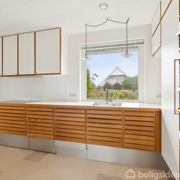 Køkkenskabe med trælamel-design hænger og står i et lyst rum. En stor vindue med udsigt til en have skaber en luftig atmosfære. En bordplade strækker sig under vinduet.