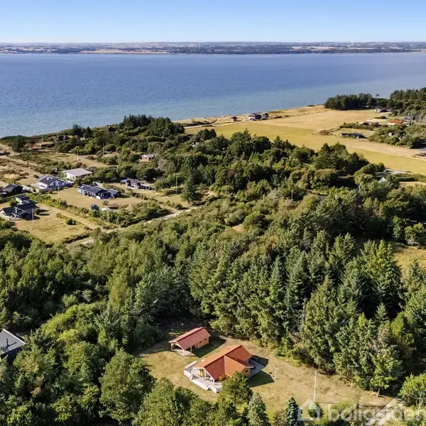 Et sommerhus omgivet af skov og mark ligger nær kysten med udsigt over havet. Området er præget af spredte bygninger og frodig vegetation.