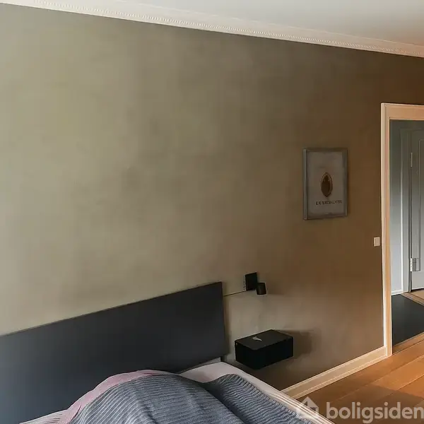 Soveværelse med en dobbeltseng dækket af et gråt sengetæppe. Et natbord og lampe er monteret på væggen ved siden af. En åben dør fører til en gang.