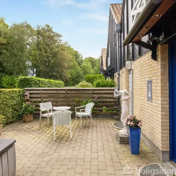 En gårdhave med fliser har et bord og stole, en klassisk statue og blomsterkrukker. Omgivet af hæk og trægærde med en blå dør på teglstensvæggen under sort udhæng.