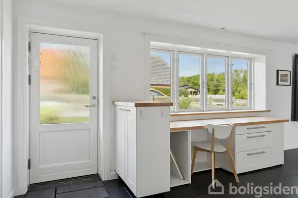 En hvid skrivebord med en stol står foran vinduer med udsigt til en grøn have. Til venstre ses en dør med mat glas, der åbner ud mod samme udsigt.