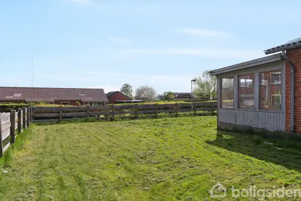Et indhegnet græsareal ligger foran en rød murstensbygning med en veranda med store vinduer; i baggrunden ses flere tage fra andre huse under en klar blå himmel.
