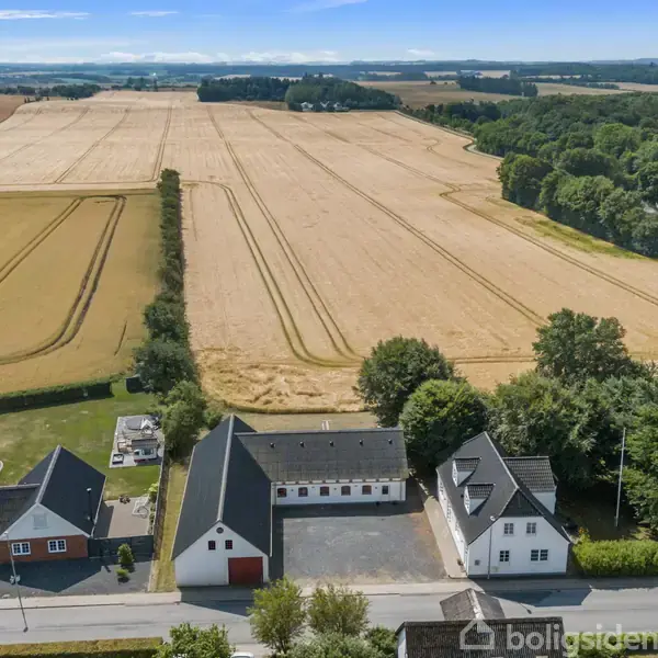 En række huse ligger langs en vej, hvoraf nogle har flade tage. Bag husene strækker store gyldne marker sig ud mod en skov, under en himmel med spredte skyer.