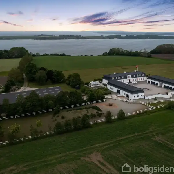 En stor hvid bondegård med staldbygninger står stille omgivet af grønne marker og træer, med en sø i baggrunden under en farverig aftenhimmel.
