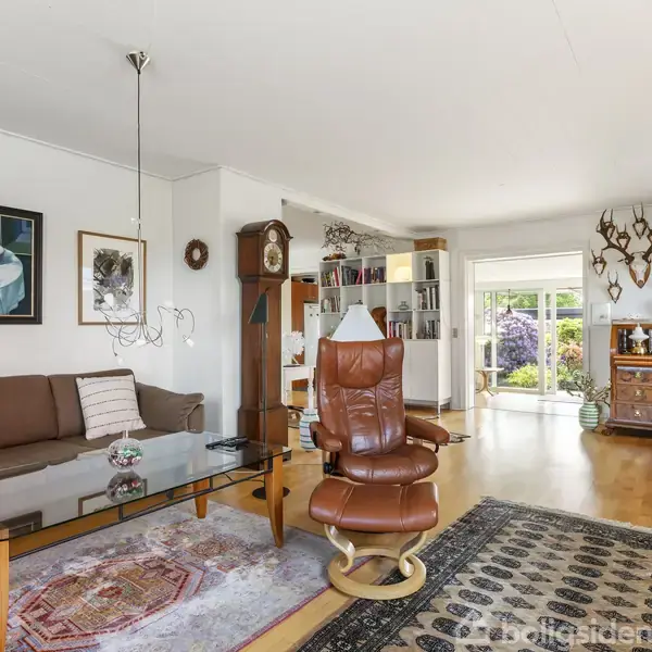 En stue med brune lædersofaer og en glasbord i midten. Vægge dekoreret med billeder og opsats. Reol og stor bogreol i baggrunden ved siden af glasskydedør mod haven.