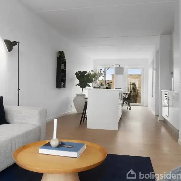 En hvid sofa står ved et rundt træbord med et stearinlys og en bog i en stue. Bagved ses et spisebord i et lyst, åbent rum med planter og køkken.