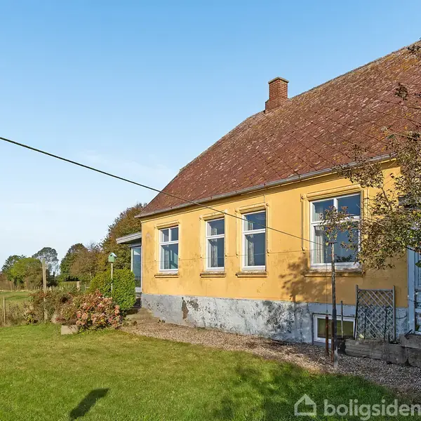 Et gult hus med rødt tegltag står i solskin, omgivet af en grøn have. I baggrunden ses åbent landskab med marker og træer.