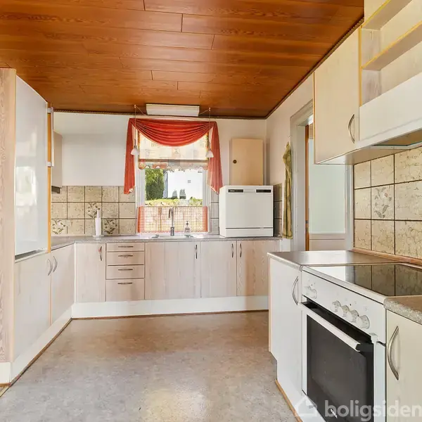 Et køkken med hvide skabe og træloft. Komfur og vask integreret i skabene. Fliser på væggen. En rød gardin over et vindue med udsigt til træer.