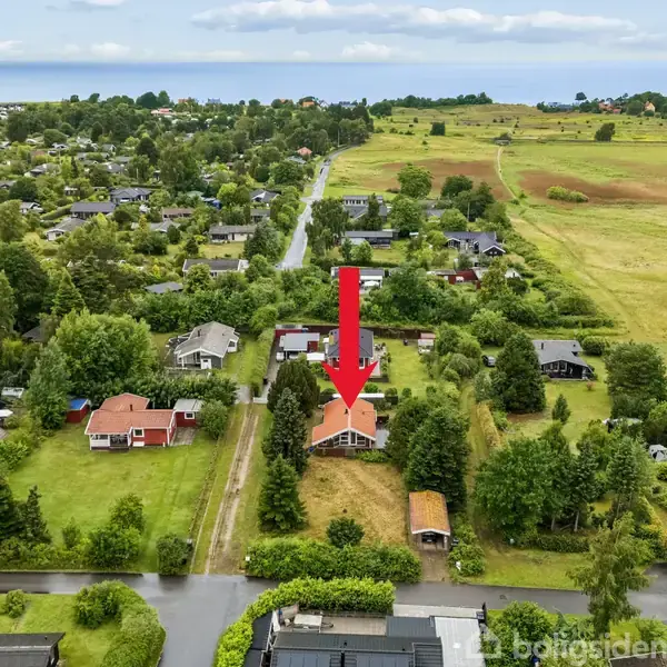Et hus i centrum er fremhævet med en rød pil. Det er omgivet af grønne træer og huse i et landligt område med marker og en fjern udsigt over havet.