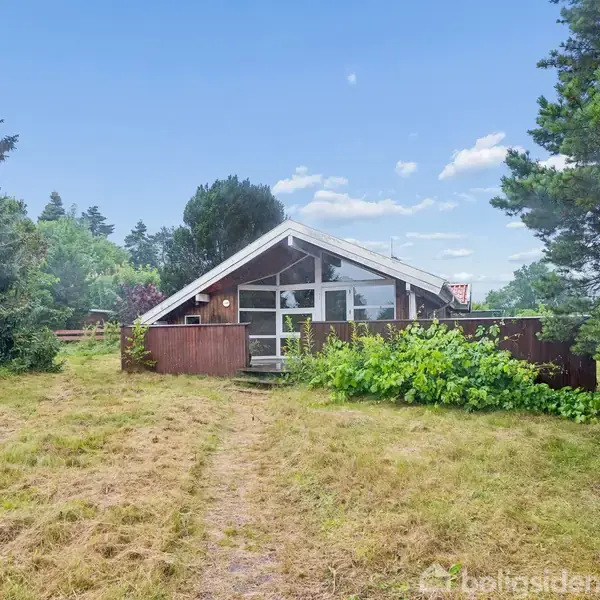 Et lille træhus med store vinduer står omgivet af højt græs og træer i en landlig have. Himlen er klar med få skyer.