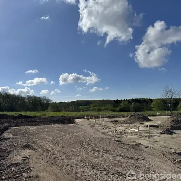 En byggeplads med jordvolde og fundamentarbejde i gang. Omgivet af grønne træer under en klar blå himmel med spredte skyer.