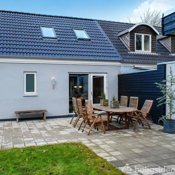 En enkelthus med mørkt tegltag og ovenlysvinduer står ved siden af en terrasse, hvor der er et bord med træstole og en udendørs grill. Græsplæne og hegn omkranser området.