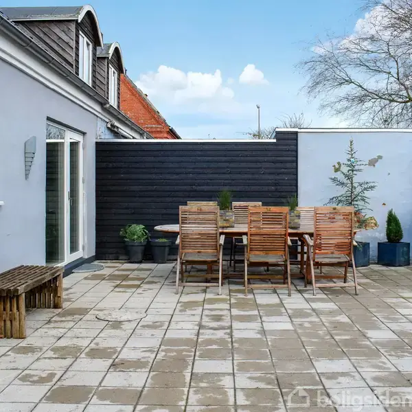 En udendørs terrasse med et træbord omgivet af syv stole står på en flisebelagt gårdsplads. Til højre ses en grill og dekorative potteplanter langs væggen.