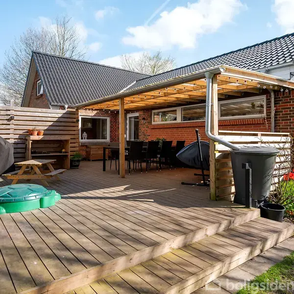 Træterrasse med borde, stole og grill står ved siden af et murstenshus. Terrasseoverdækning giver skygge. Grønt græs og blomsterbede omkranser området, mens himlen er blå med skyer.