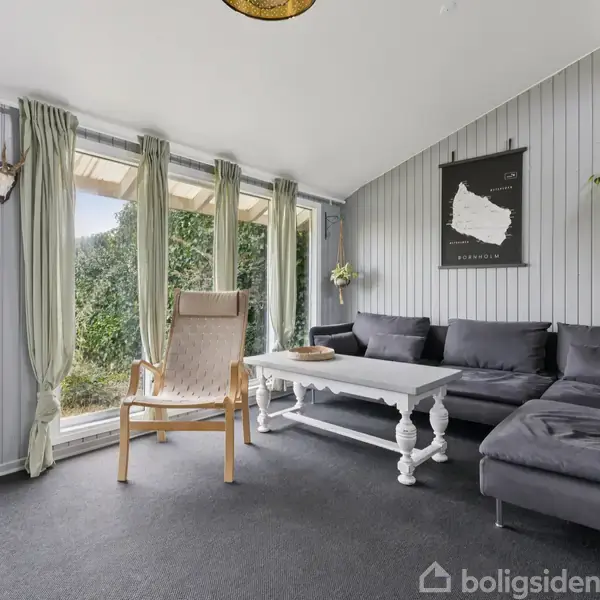 En lys stue med grå trævægge og store vinduer; en beige lænestol står ved et hvidt sofabord foran en mørkegrå sofa. På væggen hænger et kort med teksten "Anholt".