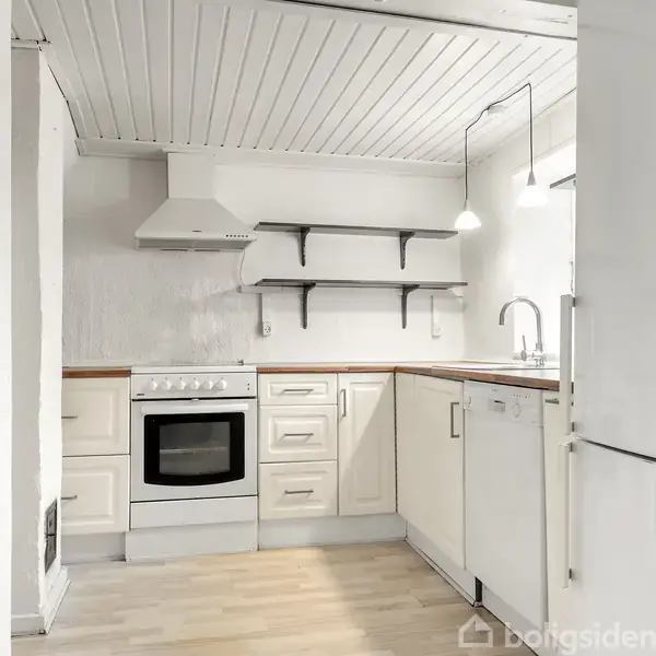 Et hvidt køkken med en ovn, hylder og vask. Lyse trægulve og hvidt træloft kompletterer rummet. En emhætte hænger over komfuret, og der er en kold hvid atmosfære.