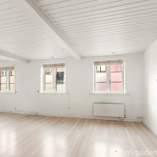 Et lyst rum med træloft har fire vinduer, der lukker dagslys ind. Rummet er omgivet af hvide vægge og lyse trægulve. En radiator er monteret ved væggen.