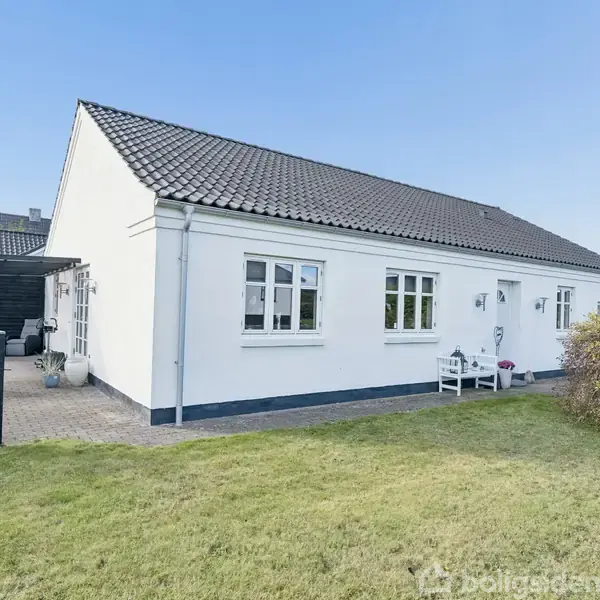 En hvid, fritliggende villa med mørkt tag. Den har en lille veranda og er omgivet af en velplejet grøn græsplæne og hæk. En terrasse ses ved siden af.