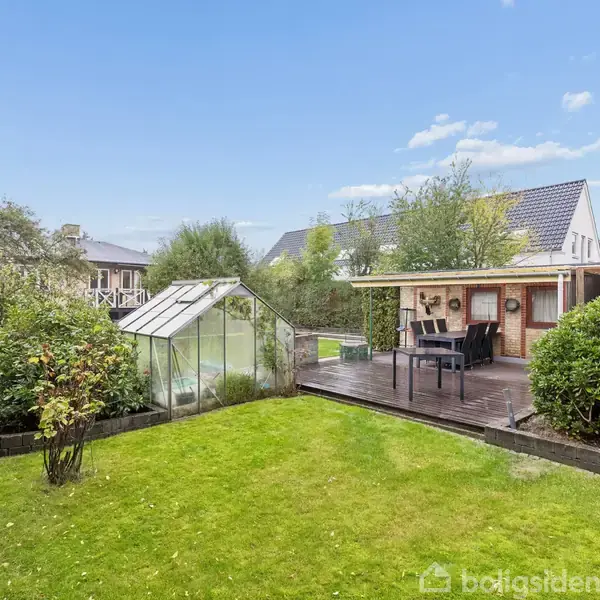 Drivhus står stille i en grøn have, mens en overdækket terrasse med bord og stole skaber en hyggelig atmosfære. Omgivet af buske og træer under en blå himmel.