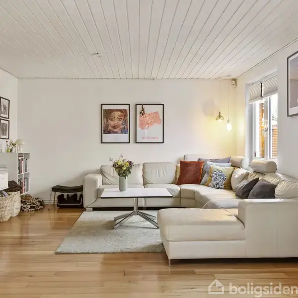 En lys stue med hvid hjørnesofa fyldt med puder, der står på et trægulv. På væggen hænger forskellige kunstplakater. Ved siden af står et fjernsyn og en kurv.