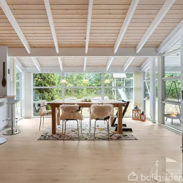 Et spisebord med stole står i et rummeligt rum med panoramavinduer, som åbner ud til en grøn have. Loftet er hvidt med synlige bjælker, og der er en åben køkkenbar til venstre.