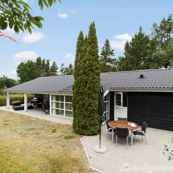 Et sort sommerhus står i en grøn have med terrasse. Et bord med fire stole er placeret på terrassen. En høj flagstang med et Dannebrog flag svajer i baggrunden.