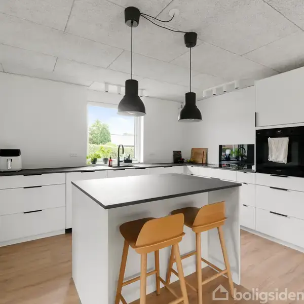 Køkken med hvidt interiør og sorte overflader, to hængelamper over køkkenø med to træbarkrakker, vindue med udsigt til have, og indbygget ovn og mikroovn i skabsmiljø.