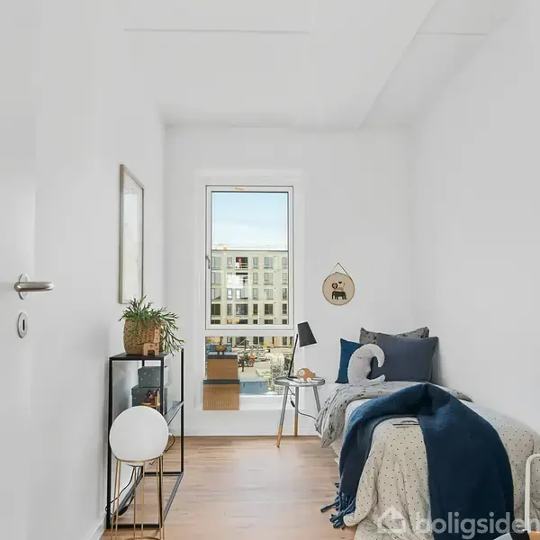 Et lille værelse med en enkeltseng fyldt med puder, en lampe på et bord ved siden af, planter på en reol, og et vindue med udsigt til en moderne bygning.