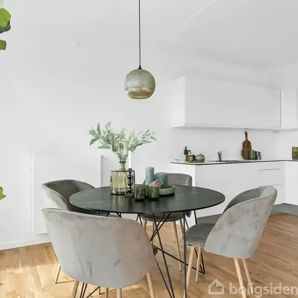 Rundt spisebord med fem grå stole står i køkkenalrum med hvide skabe og trættegulv. En grøn plante og dekorative elementer skaber en hyggelig atmosfære. En hængelampe hænger over bordet.