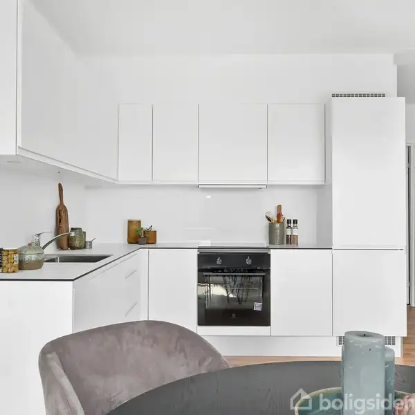 Et moderne køkken har hvide skabe og en indbygget ovn. En rund spisebordsoverflade er i forgrunden, og rummet er minimalistisk indrettet med køkkenredskaber på bordpladen.