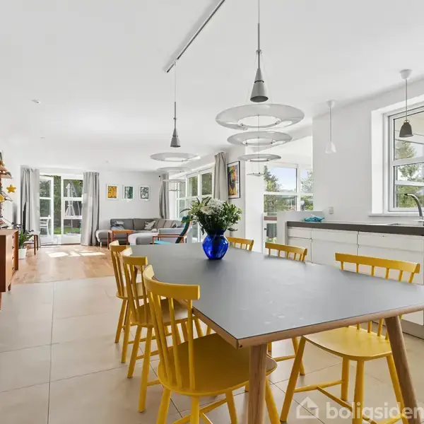 Et spisebord med gule stole står centralt i et moderne køkken-alrum. Bordet er dekoreret med en blå vase. Rummet er lyst med store vinduer og hvide vægge.