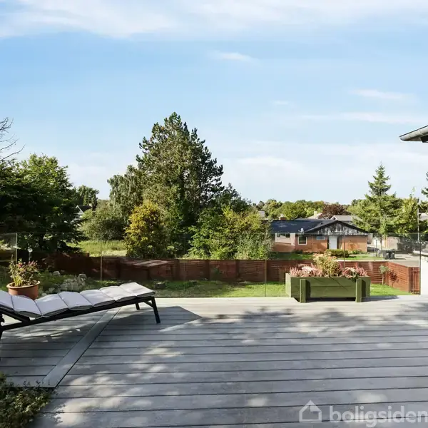 En liggestol står på en træterrasse ved siden af et glasrækværk. Omgivet af grønne træer og huse i nabolaget. Solrig himmel i baggrunden.