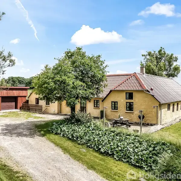 Et gult hus med rødt tegltag ligger i en grøn have. Træer og buske omgiver det, og der er en grusvej foran. En rød lade ses i baggrunden.